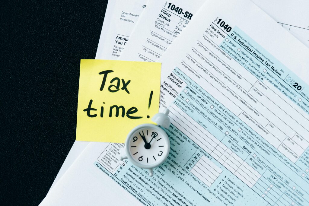 pexels-photo-6863259-6863259 A stack of tax forms with a clock and yellow sticky note saying 'Tax time!' indicating urgency.